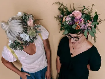 Two women wear decorative flower crowns in front of a wall.
