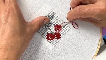 Hands cross-stitching cherry design on fabric.