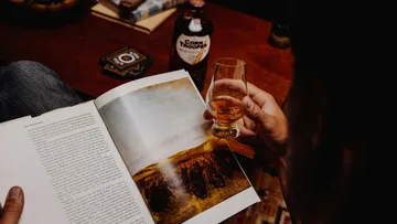 Person reading book holding whiskey glass.