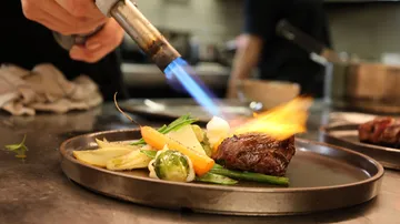 Chef torching steak with vegetables on plate.