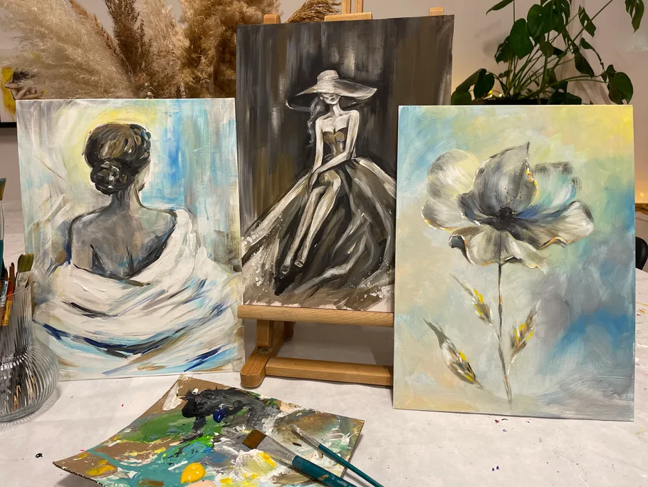 Three paintings stand on a table, surrounded by painting utensils.