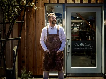 Man in apron stands in front of a restaurant with wooden façade.