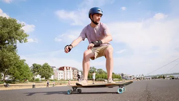 Mann fährt Skateboard auf offener Straße, Stadt im Hintergrund.