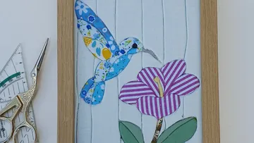 Quilted bird hovering near flower in frame.