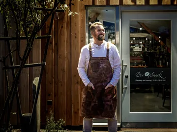 Mann im Schürze steht vor einem Restaurant mit Holzfassade.