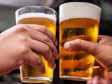 Two glasses of beer collide, hands are visible.
