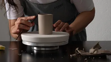 Hands sculpting clay cup on rotating stand.
