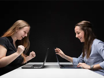 Zwei Frauen freuen sich vor Laptops an einem Tisch.