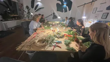 Table full of plant materials; people create Christmas decorations.
