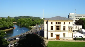 Historic building by river with trees and road.