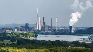 Industrieanlage emittiert Rauch nahe Fluss und Bäumen.