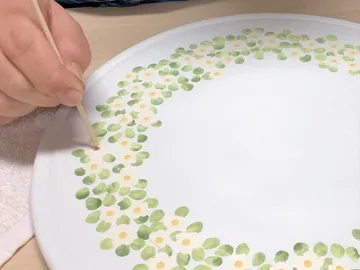 A hand paints with a pen on a white plate.
