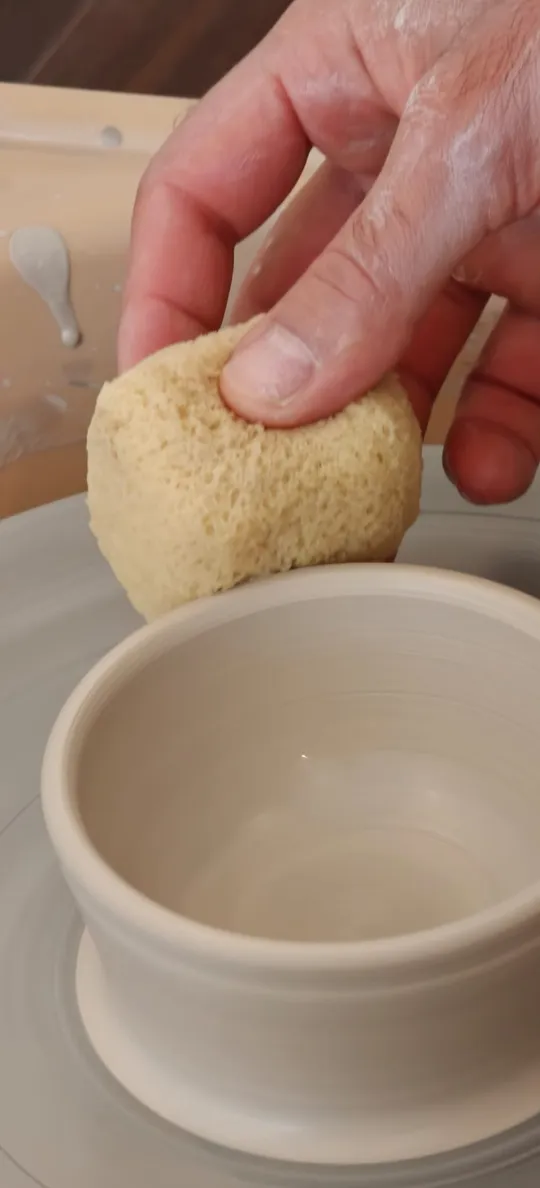 Hand schwämmt eine Keramikschale auf Drehscheibe.
