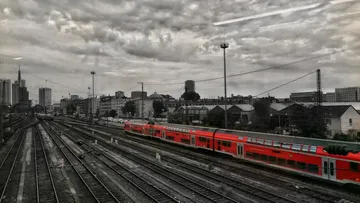 Roter Zug durchquert städtisches Bahngelände, bewölkter Himmel.