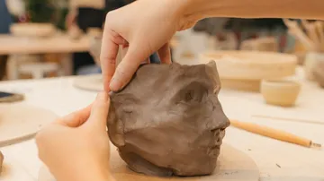 Hands sculpting a clay face on a table.
