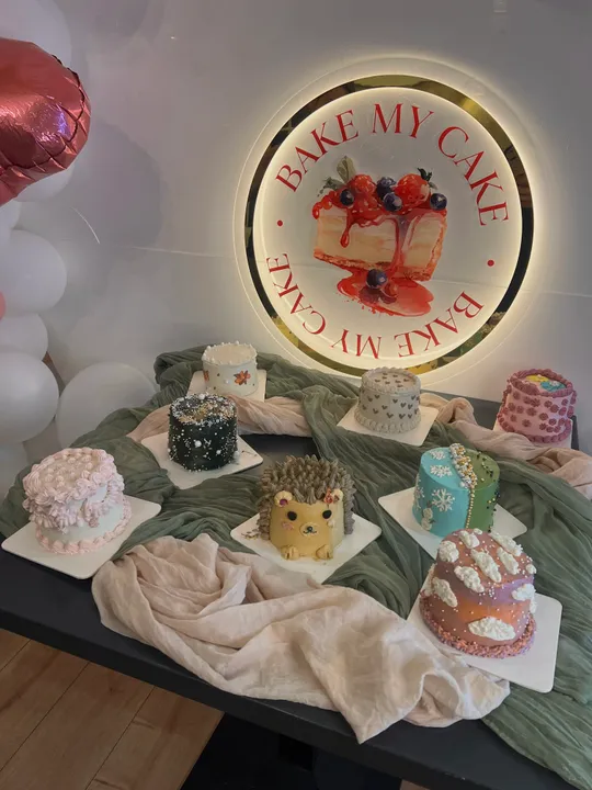 Dekorative Kuchen auf Tisch unter 'Bake My Cake' Schild.
