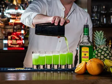 Barman pours green cocktail into glasses, fresh fruit next to it.