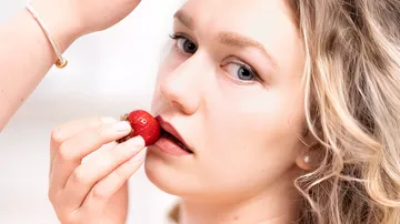 Woman holding strawberry near mouth indoors.