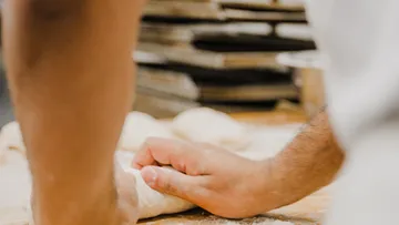 Hände kneten Teig auf bemehlter Fläche, Bäckerei im Hintergrund.