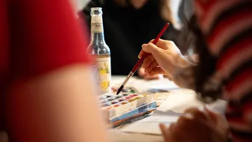 Hand holding paintbrush near watercolors, table setting.