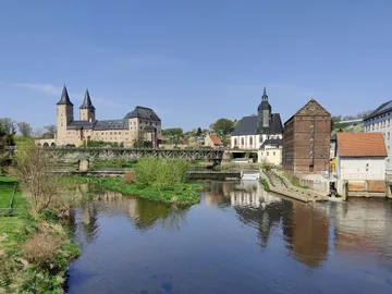 Castle and buildings reflect in a calm river under a blue sky.