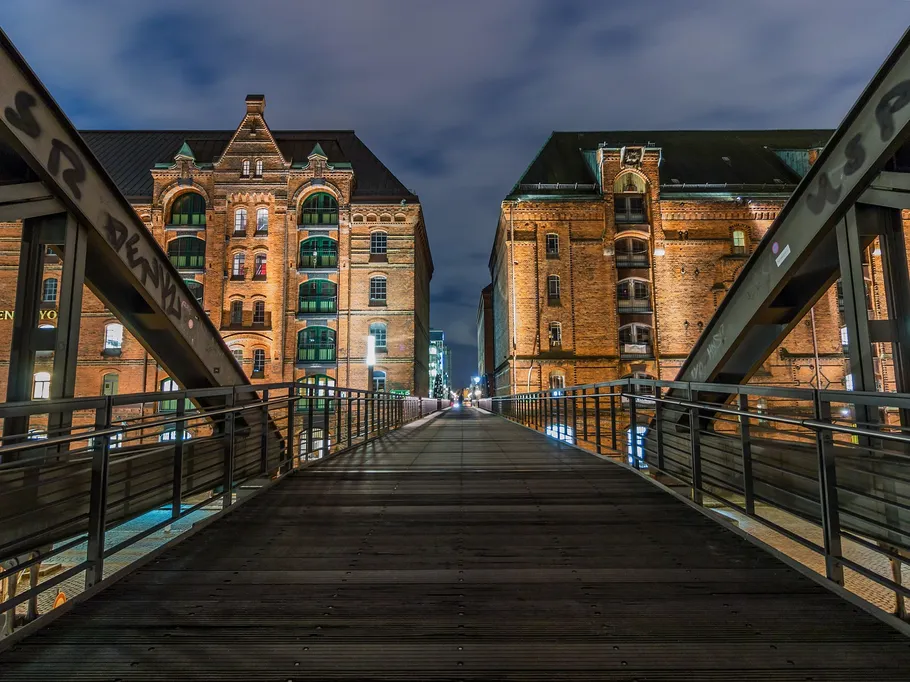 Holzbrücke verbindet historische Backsteingebäude in der Nacht.