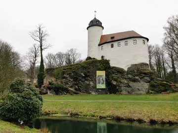 White castle stands on rocks, surrounded by trees and water.