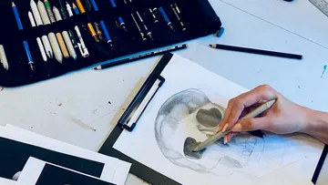 Hand sketching skull on clipboard with pencils.