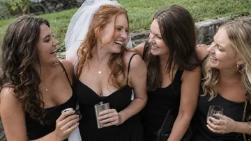 Four women laughing, wearing black dresses outdoors.