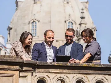 Vier Personen betrachten ein Tablet auf einem Balkon.