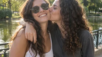 Two women smiling, one kissing the other, outdoors.