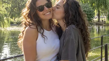 Two women smiling, one kisses the other, by lake.