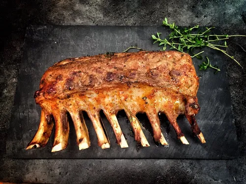 A rack of lamb lies on a black platter surrounded by thyme.