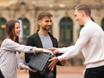 Three people hand over a black suitcase outside.