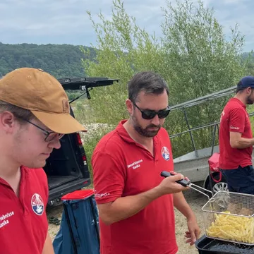Men fry food on the shore, surrounded by nature.