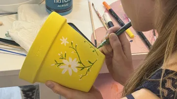 Person painting flowers on yellow pot indoors.
