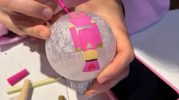 Person malt auf einem Glasornament, drinnen.