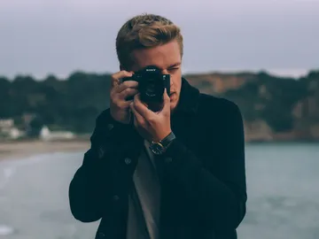Mann fotografiert am Strand mit Kamera vor Küstenlandschaft.