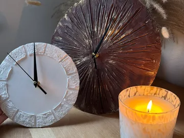 Two artistic clocks near lit candle.