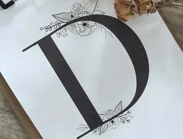 Schwarzer Buchstabe „D“ mit floralen Ornamenten auf Papier.