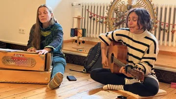 Two people playing instruments, sitting cross-legged indoors.