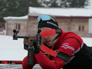 Sportler zielt mit Gewehr auf Schnee, Hintergrund mit Gebäude.