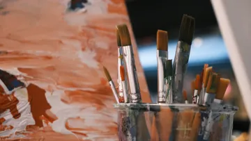 Paintbrushes in jar beside abstract canvas.