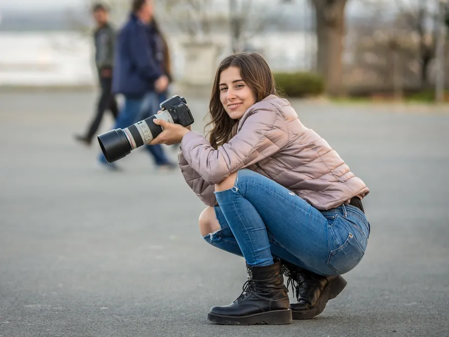 A smiling woman kneels and takes pictures outdoors.