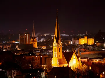 Hochhäuser leuchten nachts über einer Stadt mit Kirchen.