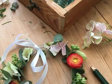 Blumenarrangement entsteht auf Holztisch, mit Schleifenband.