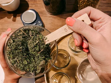 Hand holds wooden spoons with herbs over a glass container.
