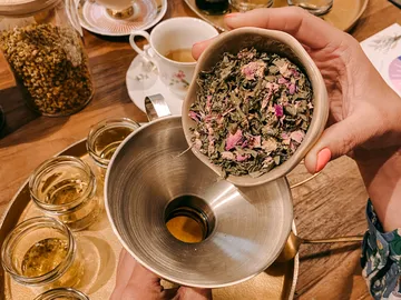 One hand holds a bowl of herbs over a funnel.
