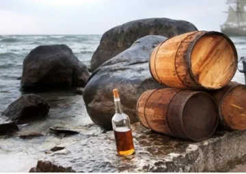 Fässer und Flasche stehen am Strand neben großen Steinen.