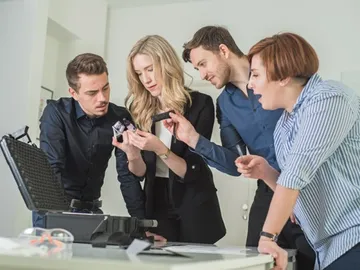 Four people examine devices on a table.